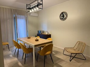 Un comedor con mesa de madera y sillas amarillas, una silla tejida, un reloj en la pared y un mueble con objetos encima.