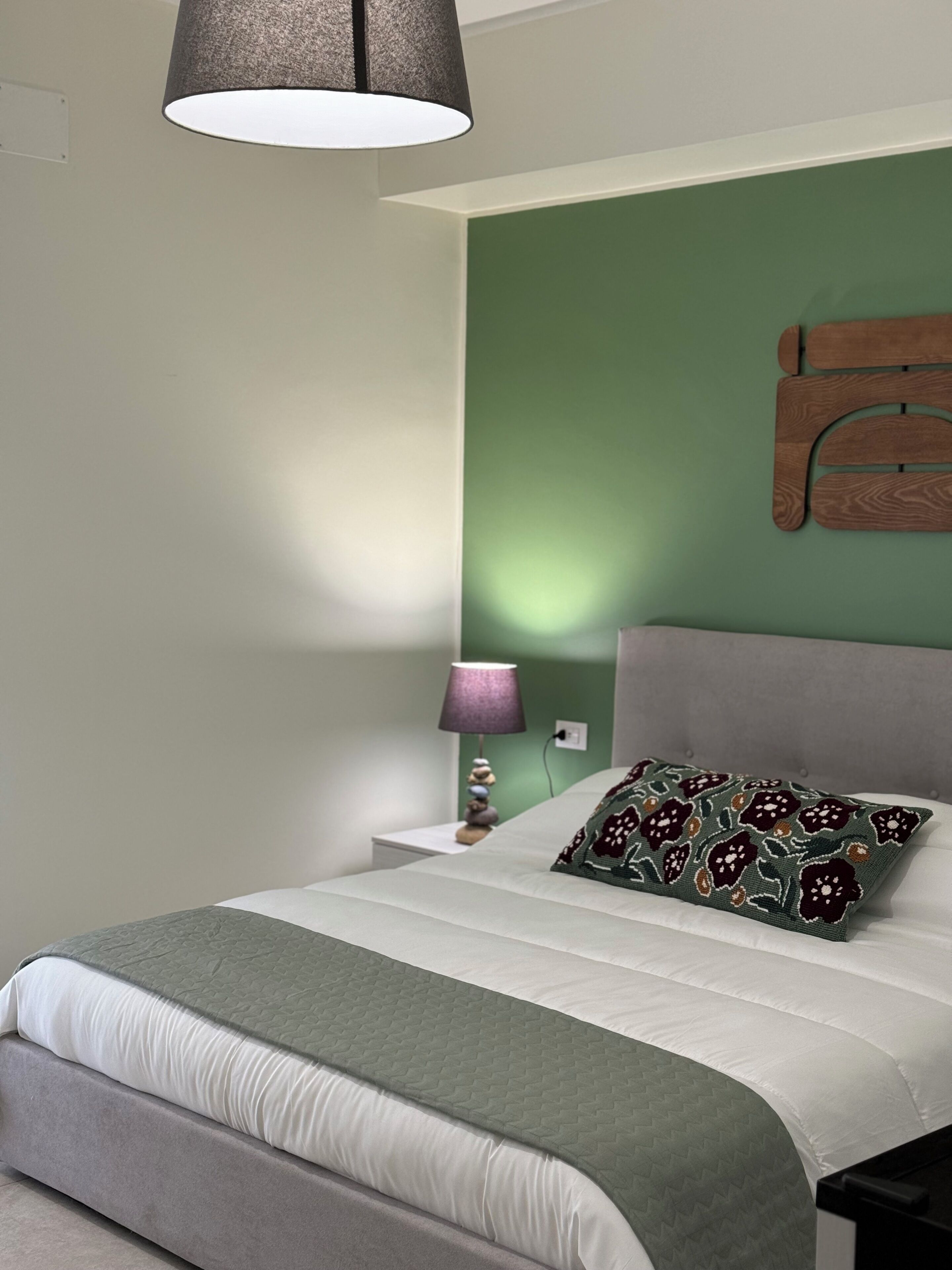 Un lit bien fait, agrémenté d’un coussin à motifs, d’une lampe de chevet et d’un objet en bois fixé au mur.