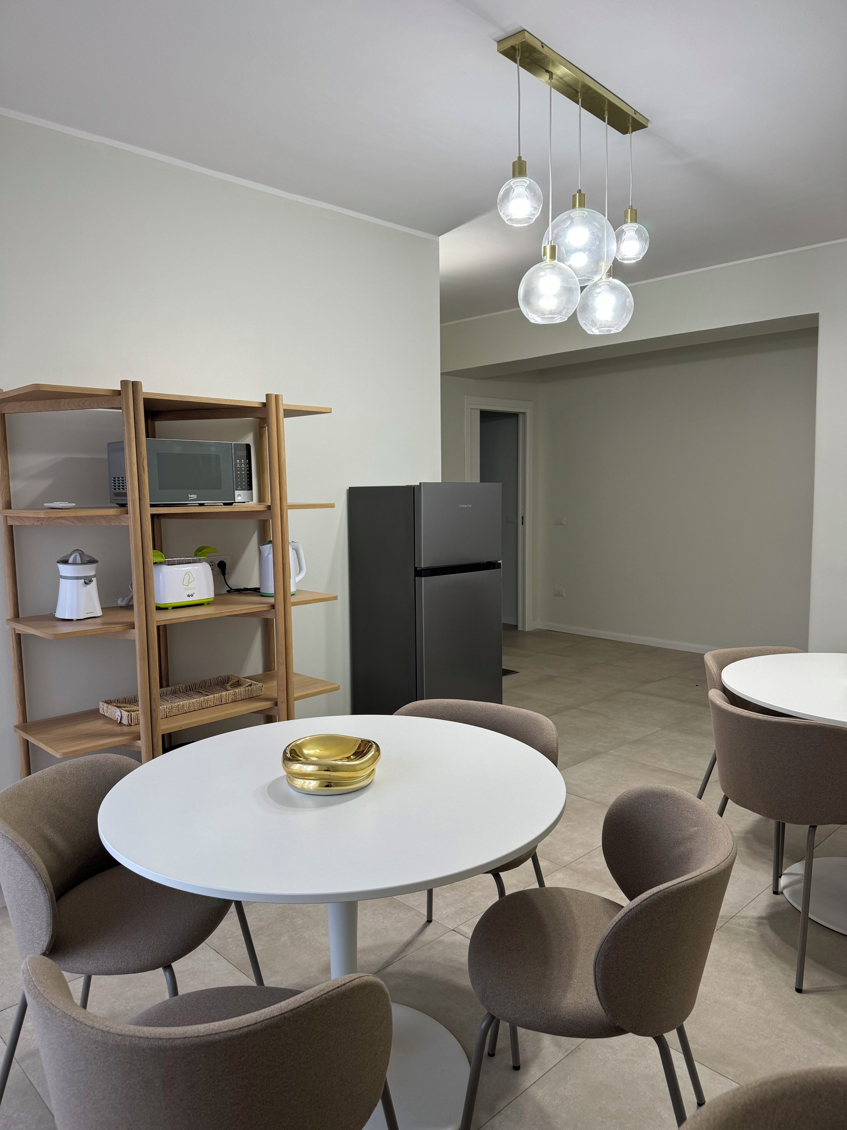A modern kitchen with a white table, brown chairs, a black refrigerator, and a wooden shelf unit with appliances.