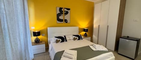 Una habitación de hotel con cama, mesitas de noche, escritorio, armario y refrigerador.