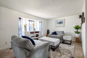 A living room with a grey sofa, a white coffee table, a grey armchair, a white side table, and a small white cabinet. There is a window with sheer curtains, a framed picture on the wall, and a potted plant on a wooden stool. - Spacious 4-bedroom house in welcoming Thornton, Cozy for families to relax. (Thornton)