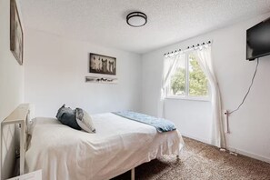 A bedroom with a bed, a window with curtains, a flat-screen TV mounted on the wall, and a ceiling light fixture. - Spacious 4-bedroom house in welcoming Thornton, Cozy for families to relax. (Thornton)