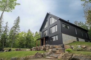 A modern two-story house with a stone staircase leading to the entrance, surrounded by a grassy area and trees. - Shack Saison - St-Lauwrence River (Petite-Rivière-Saint-François)