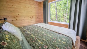 Een slaapkamer met houten wanden, een bed met bloemenbedrukking, een raam met gordijnen en uitzicht op bomen.