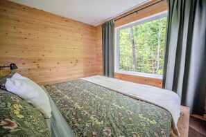 A bedroom with wooden walls, a bed with floral bedding, a window with curtains, and a view of trees. - Shack Saison - St-Lauwrence River (Petite-Rivière-Saint-François)