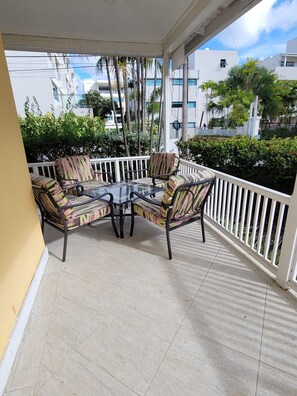 A covered outdoor seating area with a glass table and patterned chairs, surrounded by a white railing and a view of buildings and greenery. - Clariston - Traditional Barbadian home 1 minute walk from beach! (Oistins)