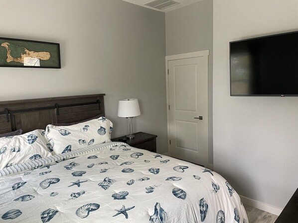 Un dormitorio con cama, mesita de noche, lámpara, puerta y televisor de pantalla plana montado en la pared.