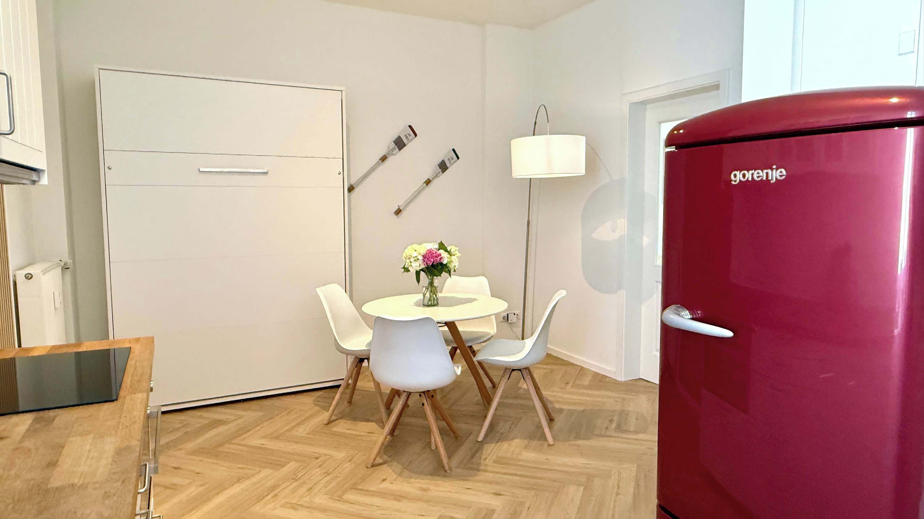 Ein kleiner Raum mit weißer Tür, einem roten Kühlschrank, einem kleinen runden Tisch mit Stühlen und einer Wandlampe.