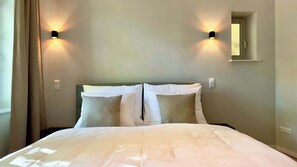 Une chambre d’hôtel avec un lit, deux lampes fixées au mur, une fenêtre avec des rideaux et un petit miroir sur le mur.