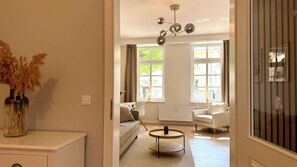 A modern living room with a sofa, a round coffee table, and a white armchair. There is a large window with curtains, a radiator, and a ceiling light fixture. - Warnemünder Auszeit 1 (Rostock)