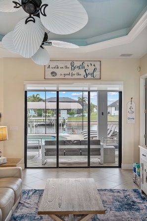 A living room with a large sliding glass door leading to a pool area, a ceiling fan, and a sign that reads 'A walk on the Beach is good for the Soul.' - Blue Tide Waterfront Pool & Hot Tub 2 En Suites (Cape Coral)