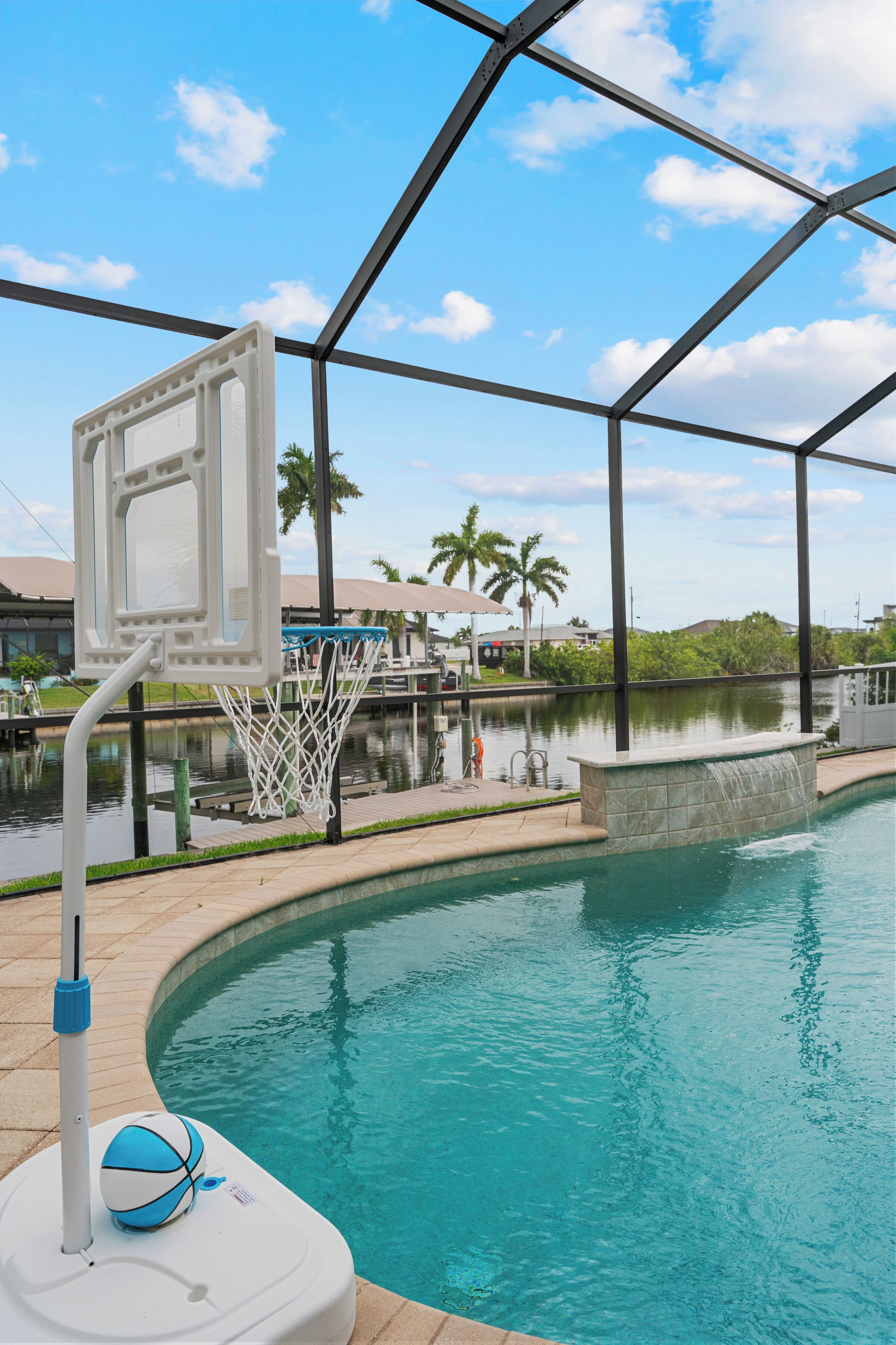 An outdoor pool with a basketball hoop, a water feature, and a view of a dock and palm trees.