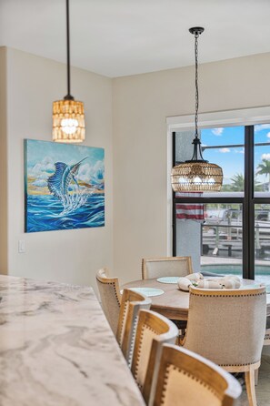 A dining area with a marble table, wicker chairs, a painting of a fish, and a view through a window. - Blue Tide Waterfront Pool & Hot Tub 2 En Suites (Cape Coral)