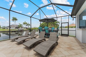 A covered outdoor area with lounge chairs, a table, and a view of a body of water and palm trees. - Blue Tide Waterfront Pool & Hot Tub 2 En Suites (Cape Coral)