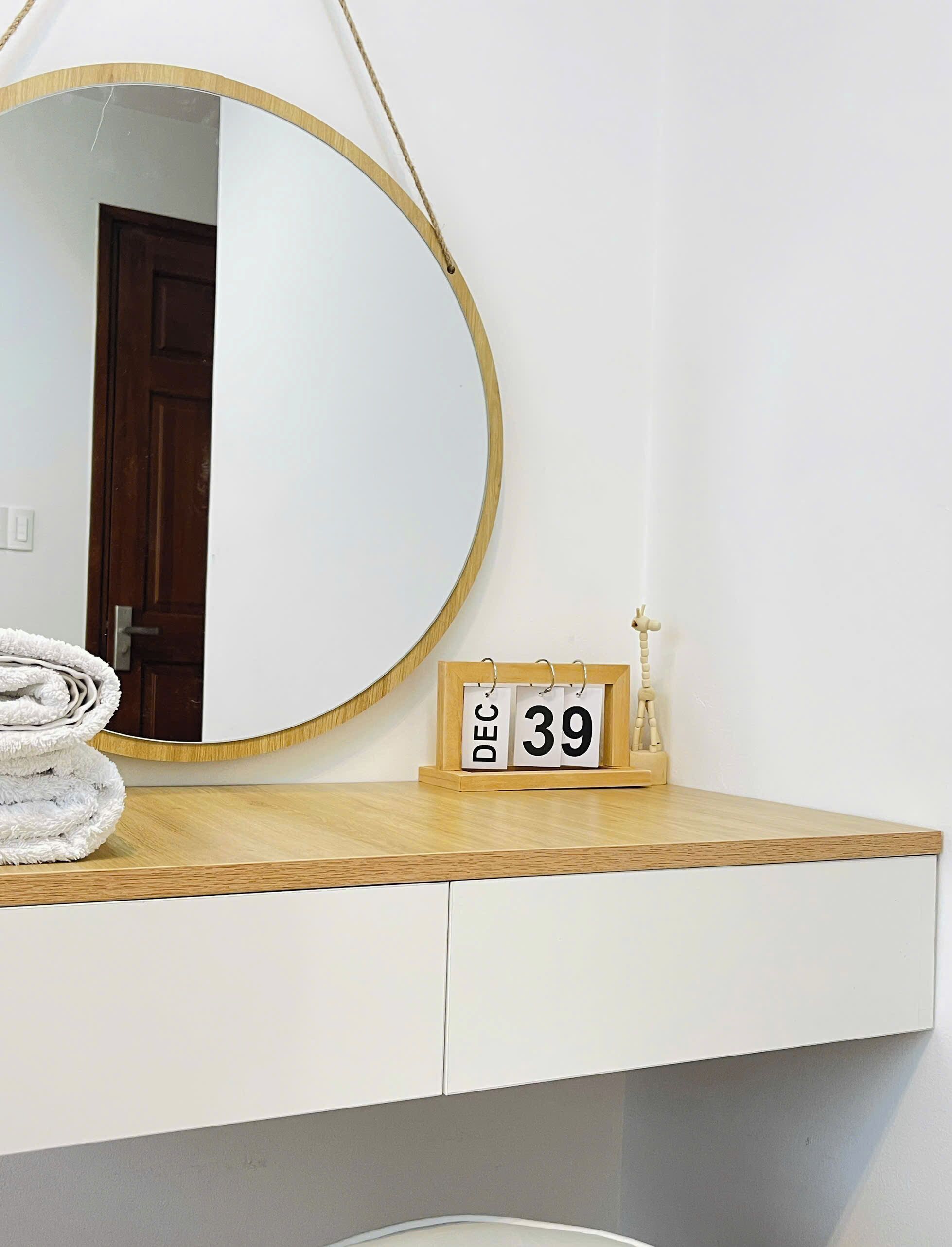 Un espejo redondo con marco de madera, un calendario de madera que muestra 'DIC 39' y una toalla blanca enrollada sobre una encimera de madera.