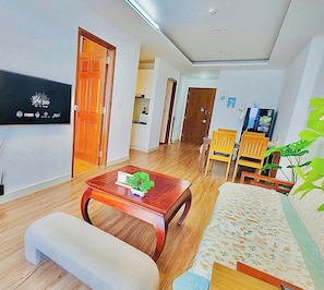 A modern living room with a sofa, a coffee table with a plant, wooden flooring, a TV mounted on the wall, and a dining area with chairs. - 2-Bedroom apartment near the airport (Ho Chi Minh City)