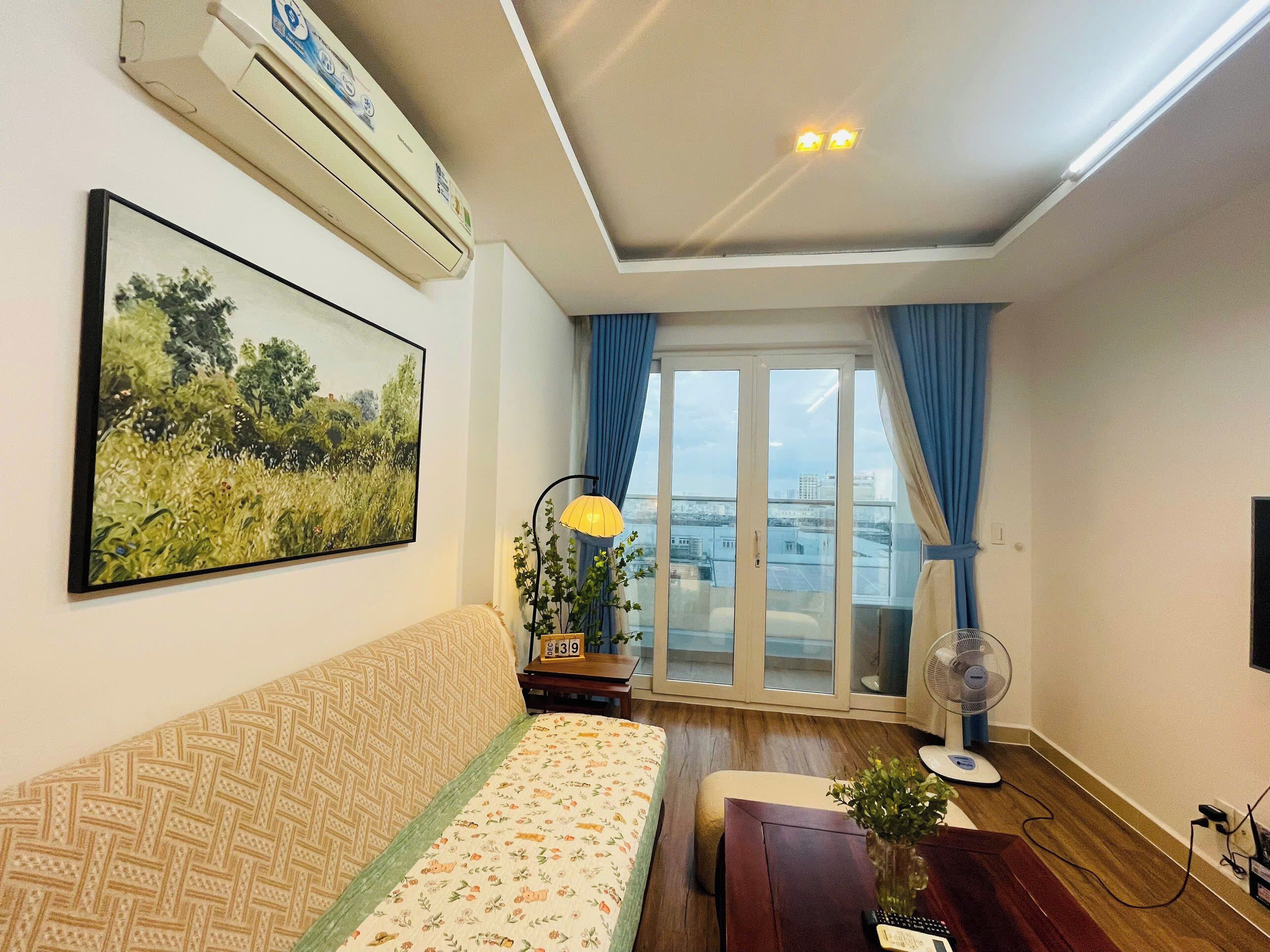 A living room with a sofa, a painting, a lamp, a fan, and a view through a glass door.