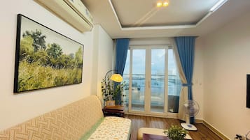 A living room with a sofa, a painting, a lamp, a fan, and a view through a glass door.