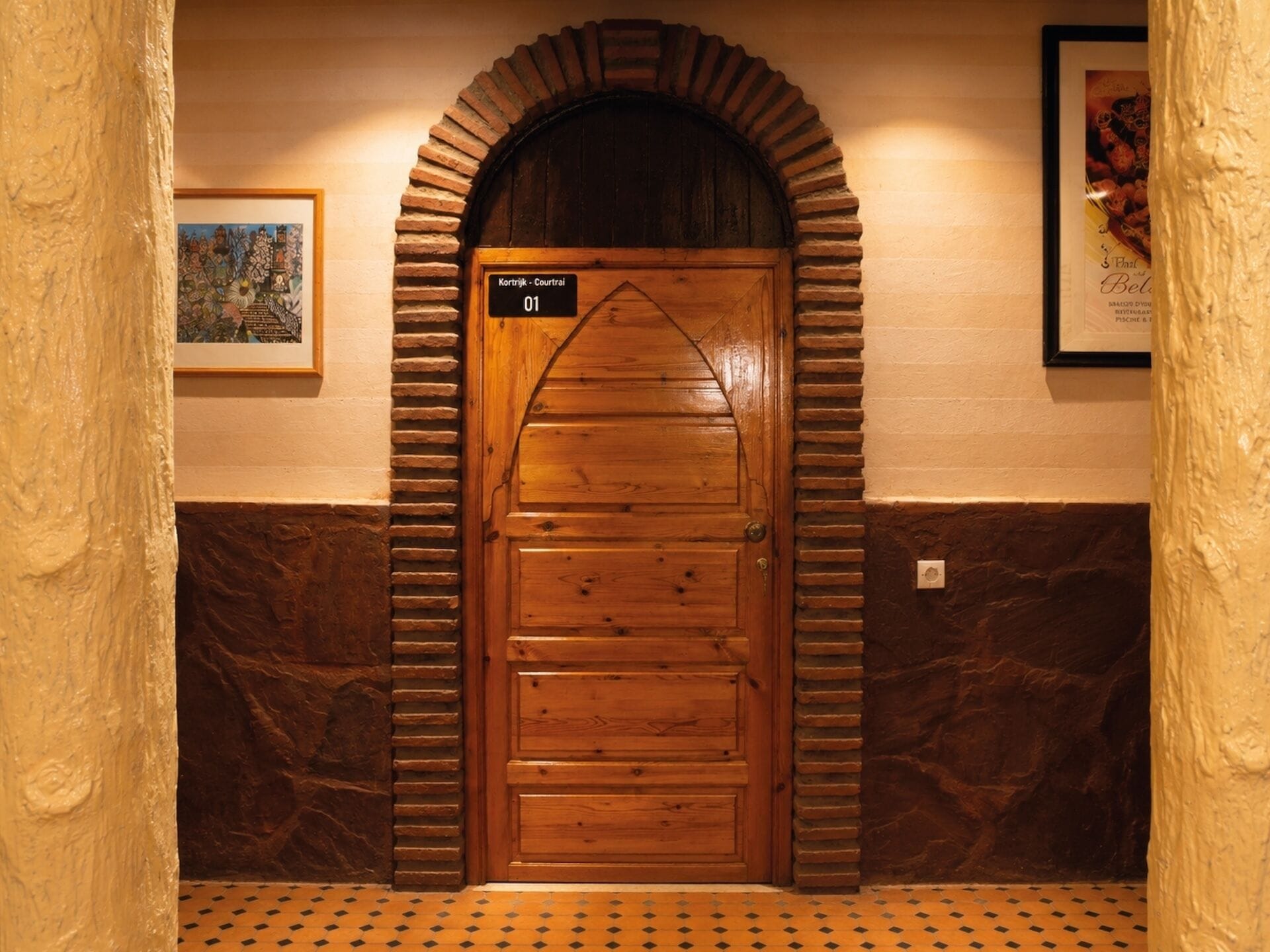 Une porte en bois avec une enseigne au-dessus, un tableau encadré accroché au mur sur la gauche et un sol carrelé.