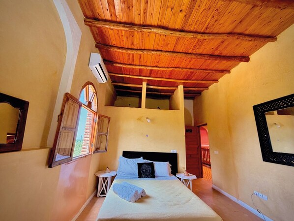 Un dormitorio con techo de madera, una cama con almohadas, una mesita de noche, una ventana con contraventanas y un espejo en la pared.