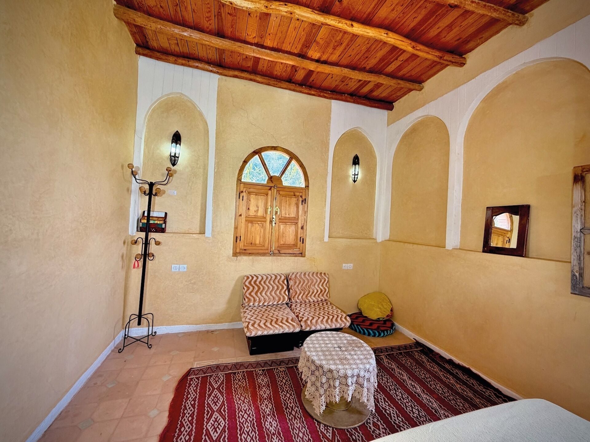 A room with a wooden ceiling, a bed with patterned bedding, a small table with a lace tablecloth, and a window with wooden shutters.