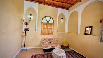 A room with a wooden ceiling, a bed with patterned bedding, a small table with a lace tablecloth, and a window with wooden shutters.