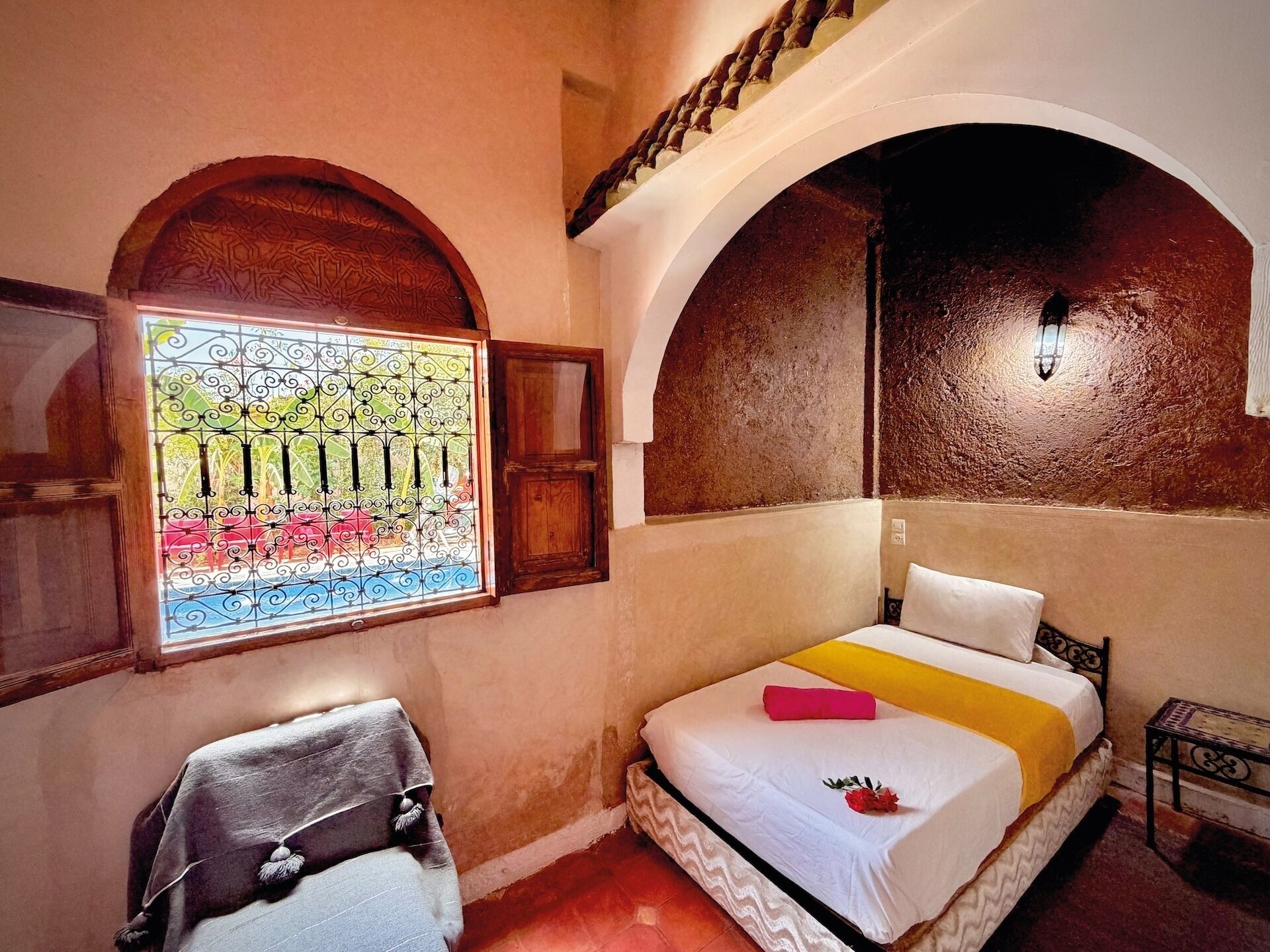 A room with a bed, a chair, a window with a view of a garden, and an arched ceiling.