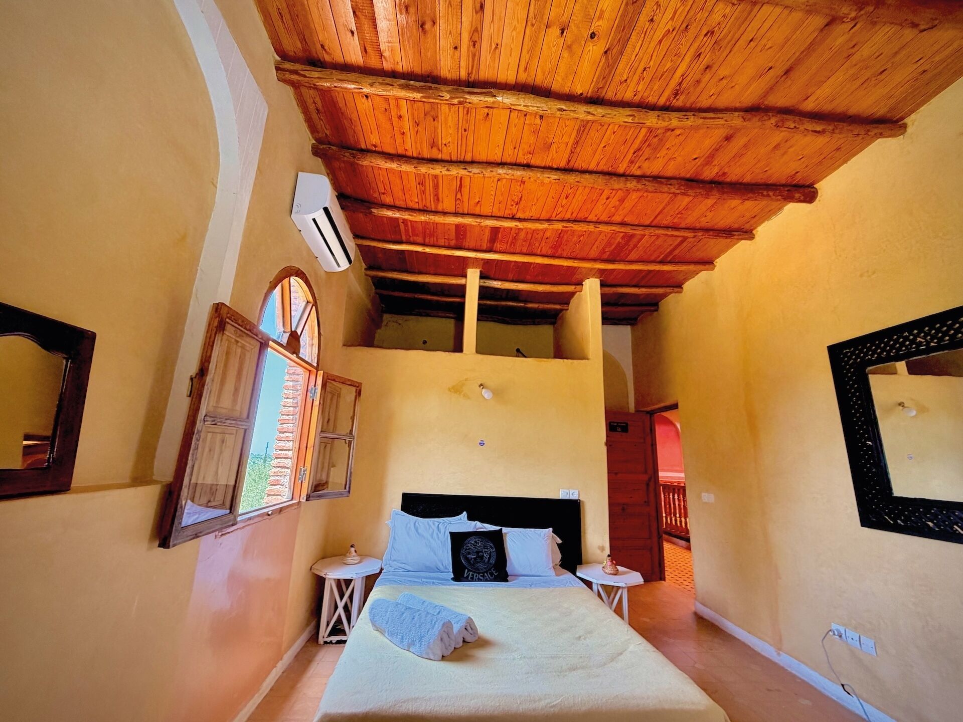 A bedroom with a wooden ceiling, a bed with pillows, a nightstand, a window with shutters, and a mirror on the wall.