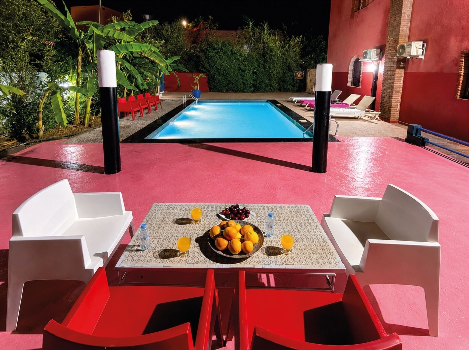 A poolside area with a table set for two, surrounded by red chairs, and a pool in the background.