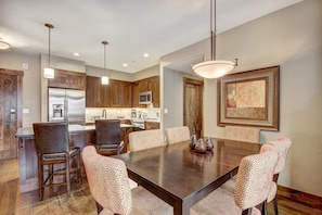 A modern kitchen with wooden cabinets, a stainless steel refrigerator, and a dining area with a large table and patterned chairs. - Waterhouse #5109 in Columbine Rd (Columbine Rd)