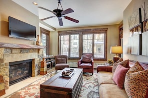 A living room with a fireplace, a television mounted on the wall, a ceiling fan, and a large window with a view of buildings outside. - Waterhouse #5109 in Columbine Rd (Columbine Rd)