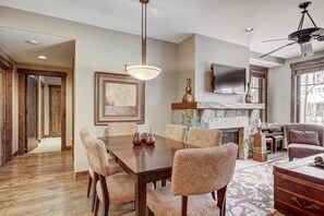 A dining area with a wooden table and patterned chairs, a fireplace with decorative items, and a television mounted above it. - Waterhouse #5109 in Columbine Rd (Columbine Rd)
