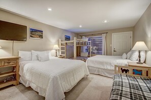 Une chambre avec deux lits, un lit superposé, une table de chevet en bois, une lampe, un téléviseur et une porte.