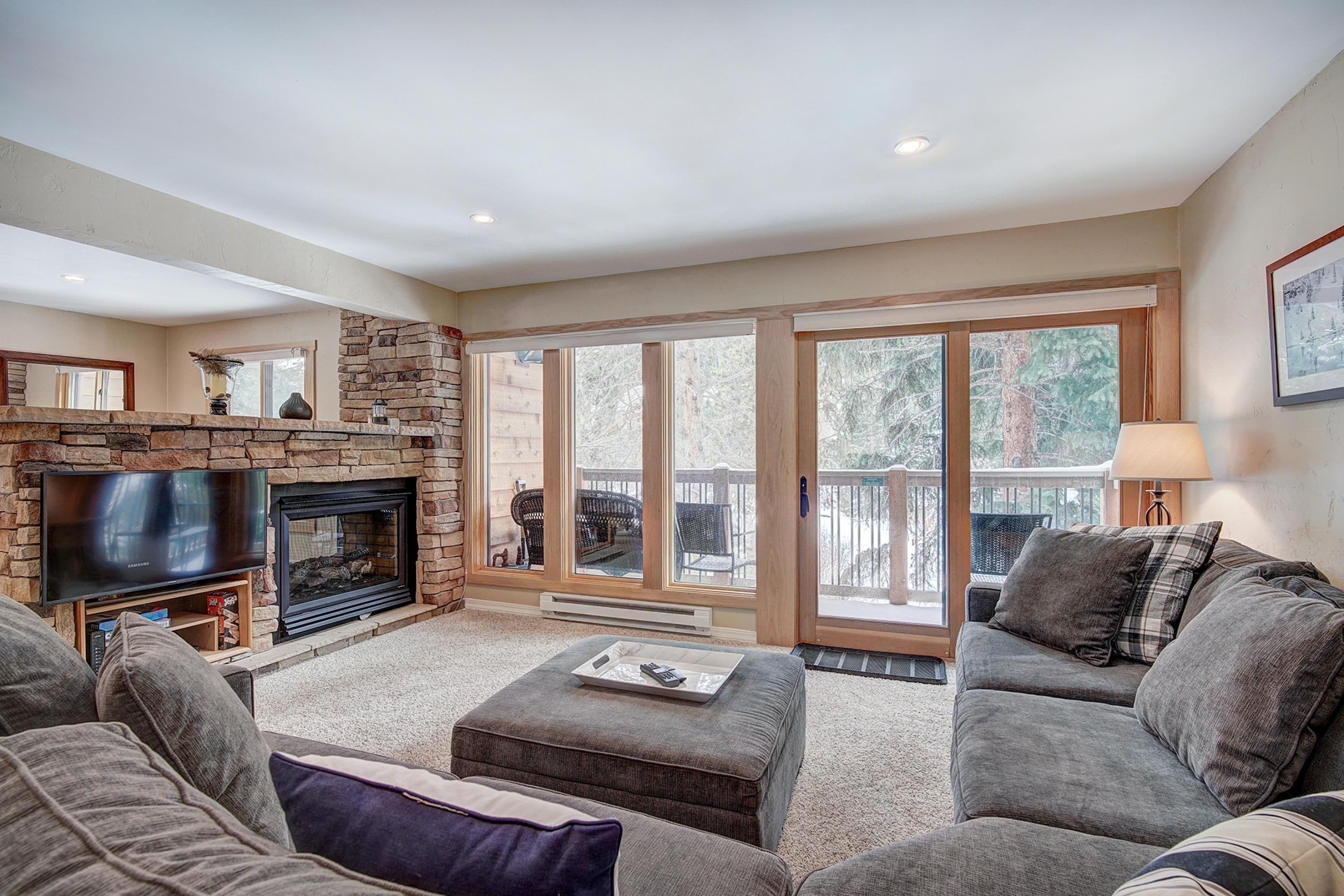 A living room with a stone fireplace, a television, and a large window overlooking a deck.
