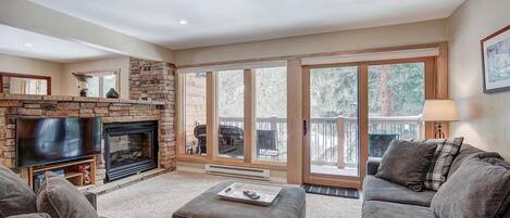 A living room with a stone fireplace, a television, and a large window overlooking a deck.