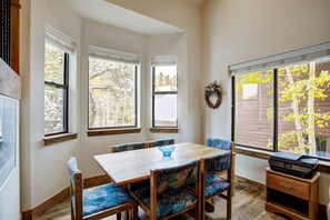 A dining area with a wooden table and chairs, large windows with views of trees, and a small wooden cabinet with a printer. - Village Point 308 in Breckenridge (Breckenridge)