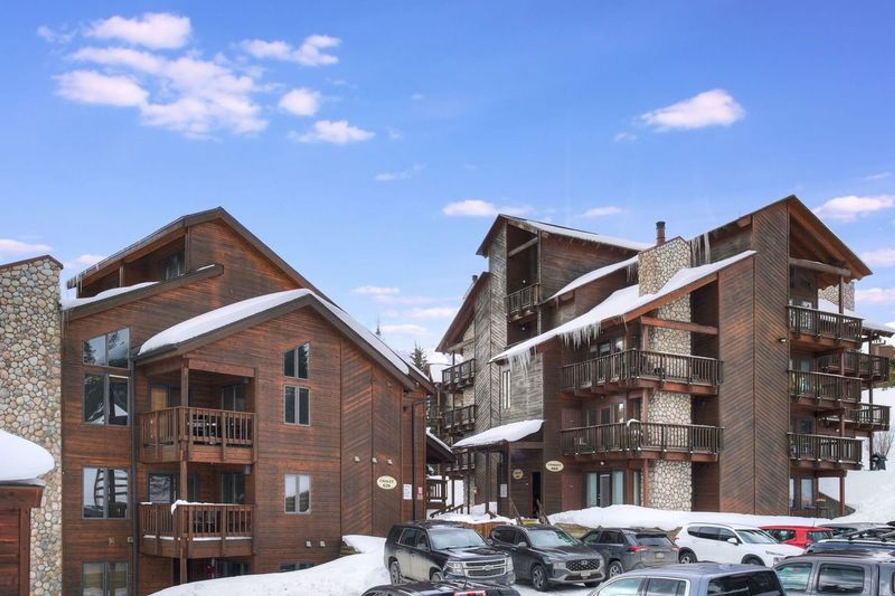 A multi-story wooden building with balconies, a stone facade, and a parking lot with cars.