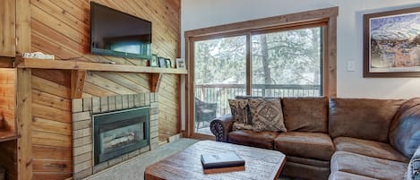 A cozy living room with a fireplace, a leather sofa, a coffee table, and a large window with a view of trees.