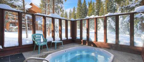 A heated outdoor pool area with a wooden deck, two chairs, and a view of a snowy forest.