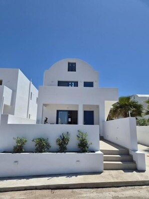 A modern white building with a flat roof, a central entrance, and a staircase leading to the entrance. - White Wave Houses (Santorini)