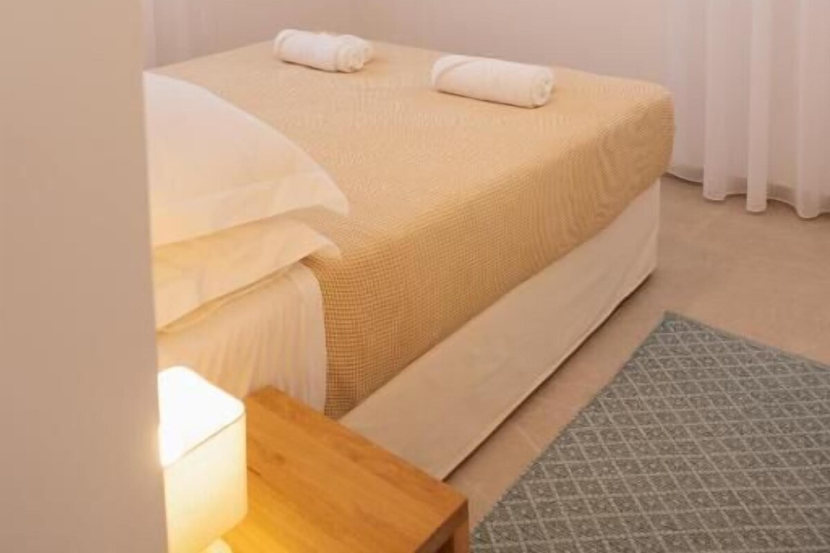 A bedroom with a bed, a flat-screen TV, an air conditioning unit, and a window with curtains.