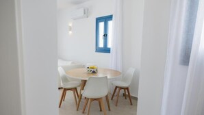 A dining area with a round table, white chairs, a bowl of fruit, and a blue-framed window. - White Wave Houses (Santorini)