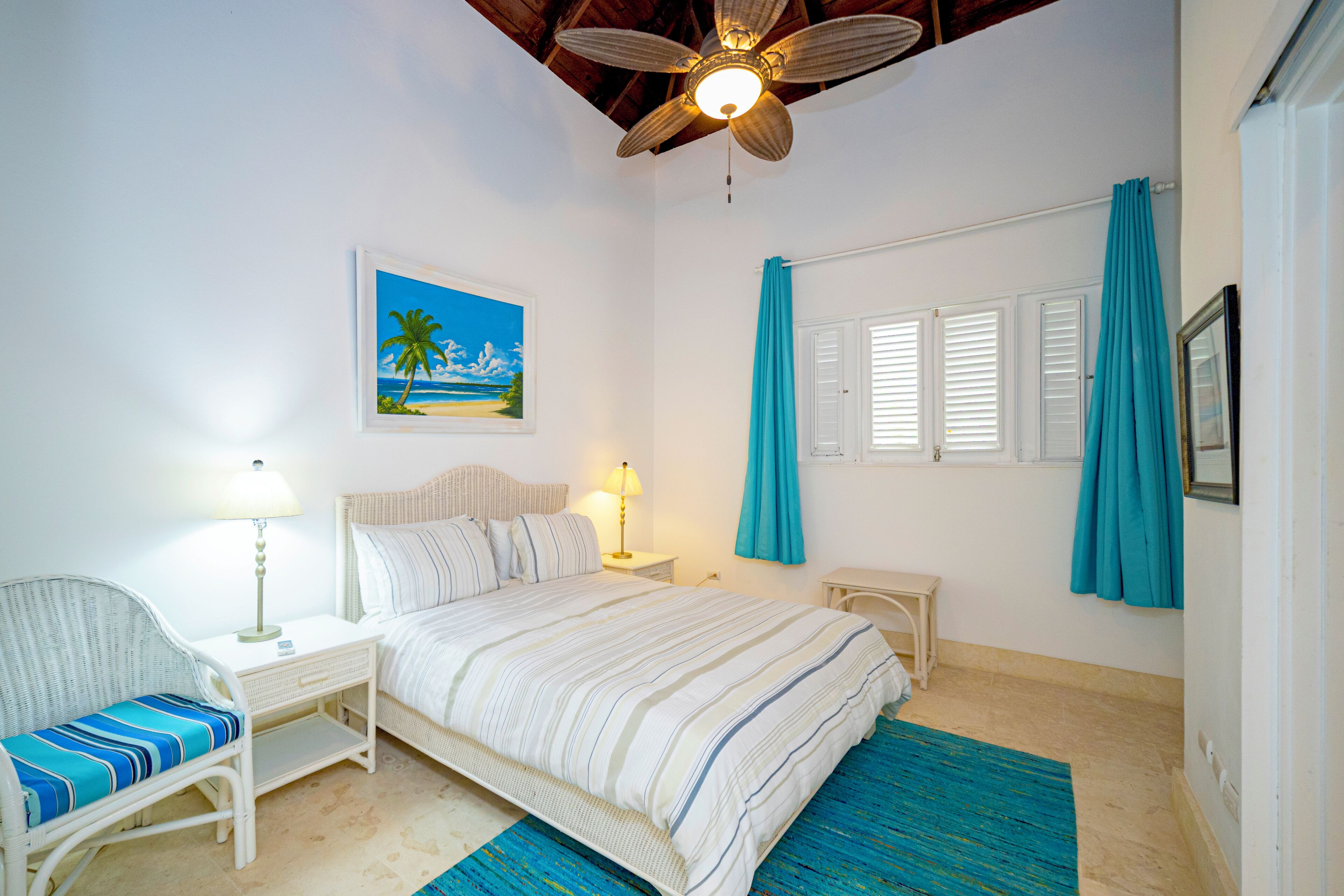 A bedroom with a bed, a chair, a nightstand, a ceiling fan, a window with shutters, and a painting on the wall.