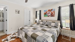 A bedroom with a bed, a ceiling fan, a painting, a window with curtains, and a door. - Sizzlin Farmhouse in Carencro (Lafayette Parish)