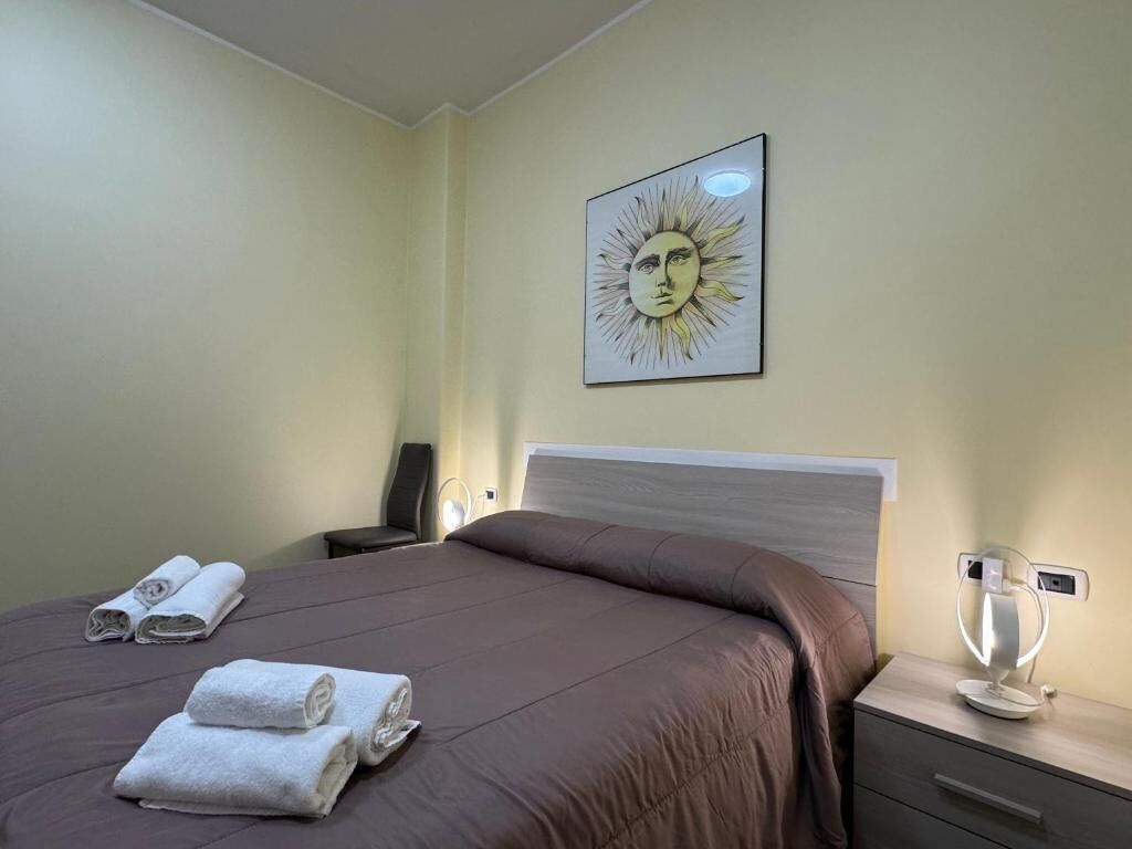 A hotel room with a bed, a bedside table with a lamp, a wall-mounted artwork, and rolled towels.