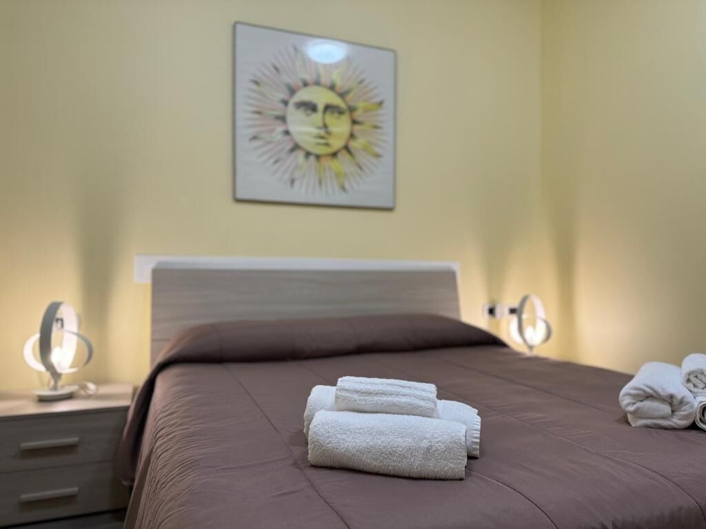Ein ordentlich bezogenes Bett mit einem braunen Kopfteil, einem an der Wand hängenden Kunstwerk einer Sonne und zwei Nachttischen mit Lampen.