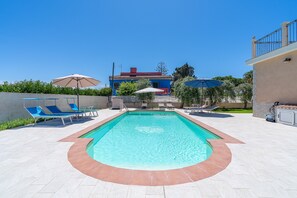 Ein Pool mit blauen Liegestühlen, ein Gebäude mit rotem Dach im Hintergrund und ein klarer, blauer Himmel.