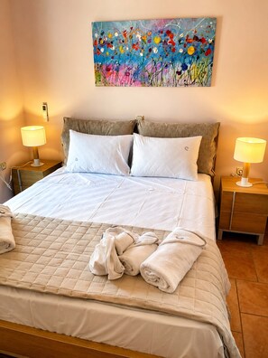 A neatly made bed with white linens, two bedside lamps, a painting on the wall, and a wooden headboard. - Suite Sunset, Overlooking the Sea in Ag. Vasilios (Agios Vasilios)