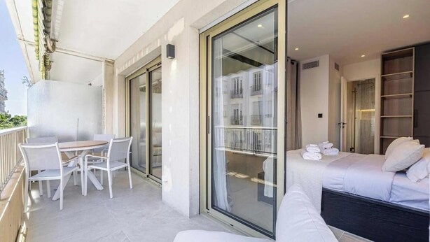 En balkon med et bord og stole, en sofa og en skydedør, der fører til et rum med en seng og et skab.