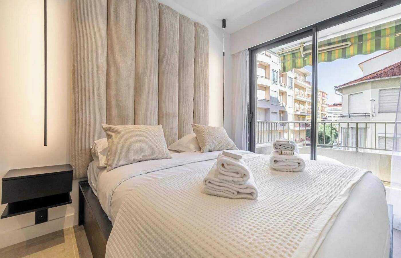 Une chambre d’hôtel avec un grand lit, une table de chevet, une fenêtre donnant sur des immeubles et un balcon.